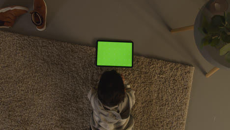 Luftaufnahme-Eines-Kleinen-Jungen,-Der-Zu-Hause-Auf-Einem-Teppich-Liegt-Und-Spiele-Spielt-Oder-Nachts-Auf-Ein-Digitales-Tablet-Mit-Grünem-Bildschirm-Streamt